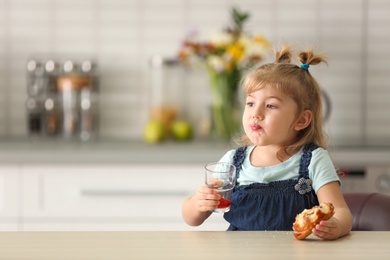 Photo of Cute little girl drinking juice with bun in kitchen