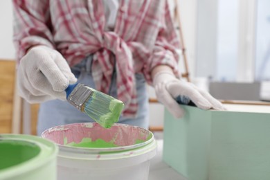 Photo of Woman dipping brush into bucket of green paint at table indoors, closeup