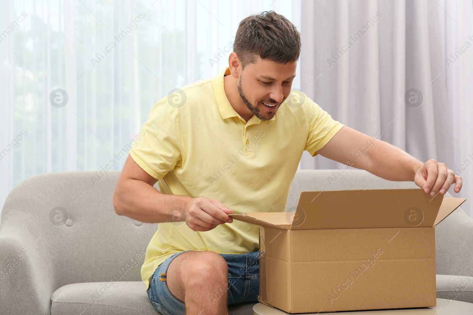 Photo of Happy man opening parcel on sofa at home. Internet shopping