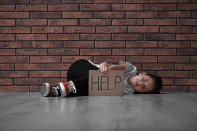 Sad little girl with sign HELP lying on floor near brick wall. Child in danger