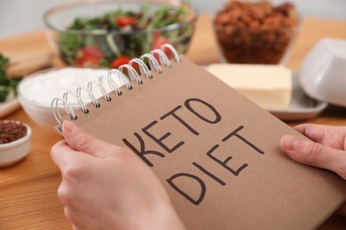 Photo of Woman holding notebook with phrase Keto Diet at wooden table, closeup