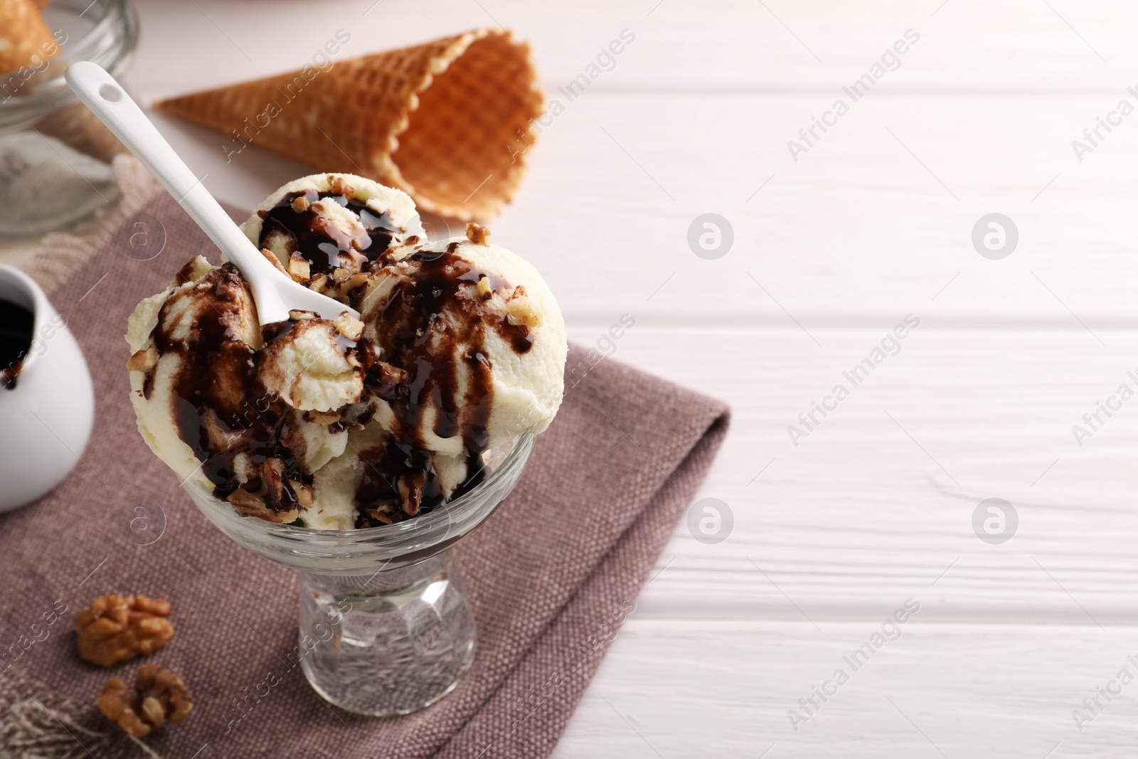 Photo of Glass dessert bowl of tasty ice cream with chocolate topping and nuts served on white wooden table. Space for text