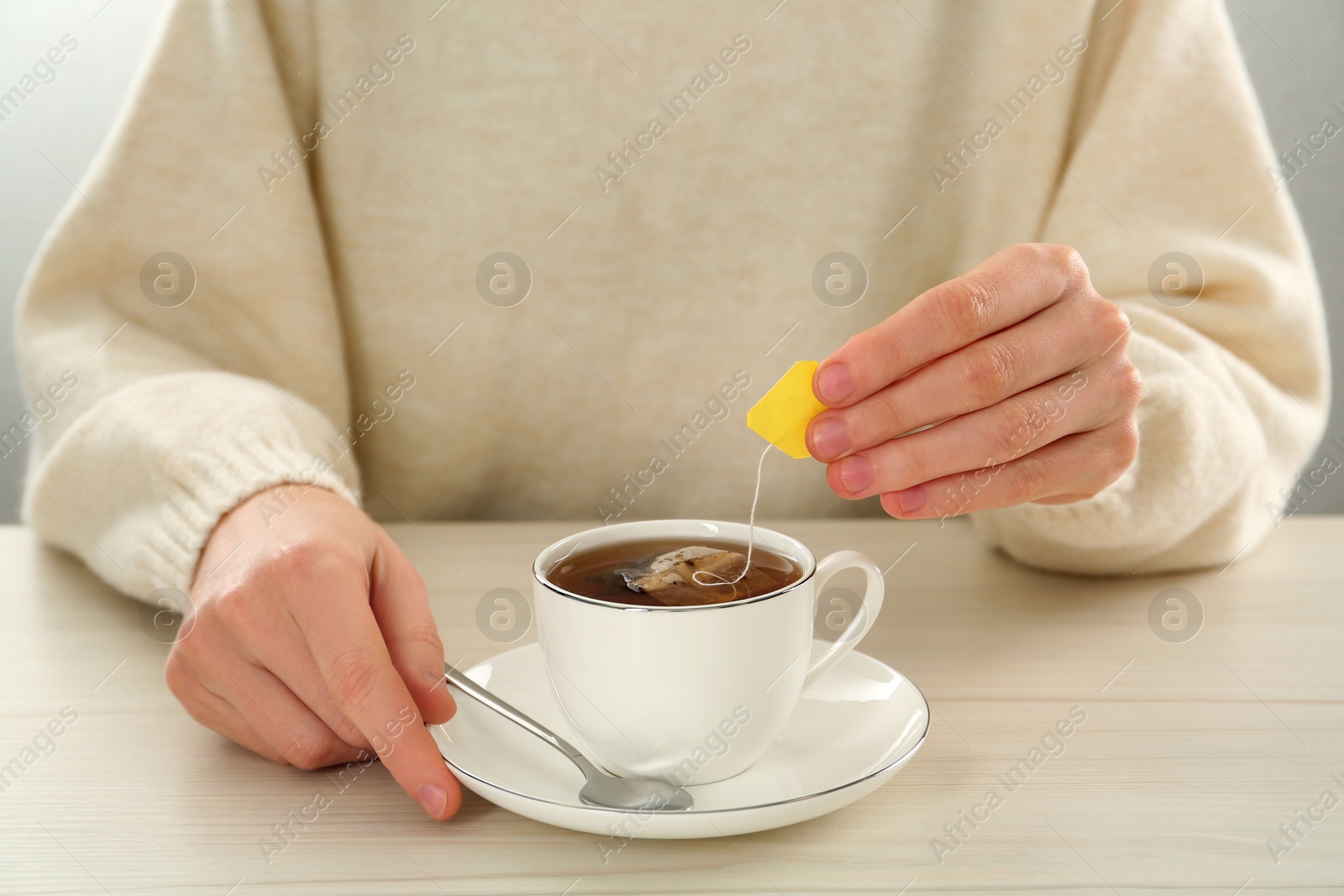 Photo of Woman dipping tea bag into cup of water at white wooden table, closeup