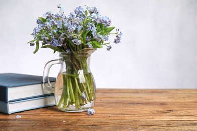 Bouquet of beautiful forget-me-not flowers in glass jug and books on wooden table against light background, space for text