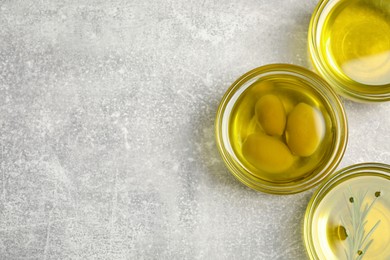 Bowls with cooking oil, olives and rosemary on light gray textured table, flat lay. Space for text