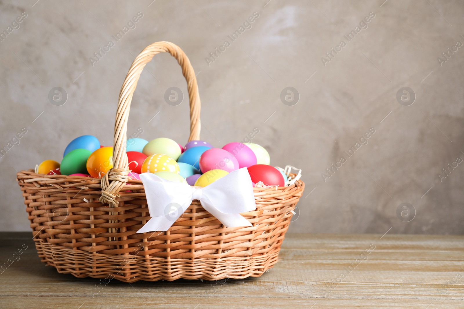 Photo of Wicker basket with bright painted Easter eggs on wooden table against grey background. Space for text