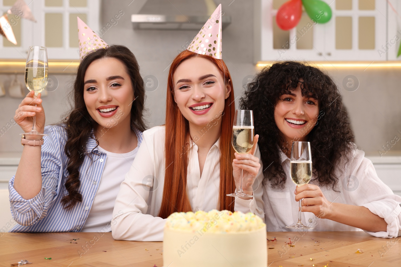 Photo of Happy young women with tasty cake and glasses of sparkling wine celebrating birthday in kitchen