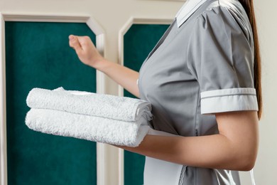 Photo of Young chambermaid with towels knocking on room door in hotel, closeup