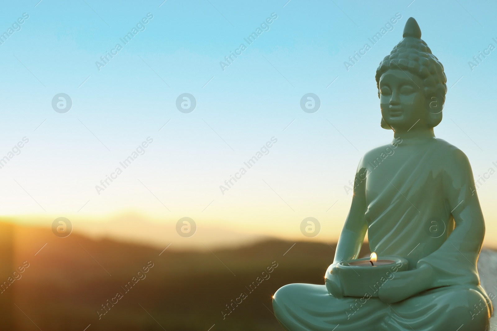 Photo of Decorative Buddha statue with burning candle on log in mountains at sunset. Space for text