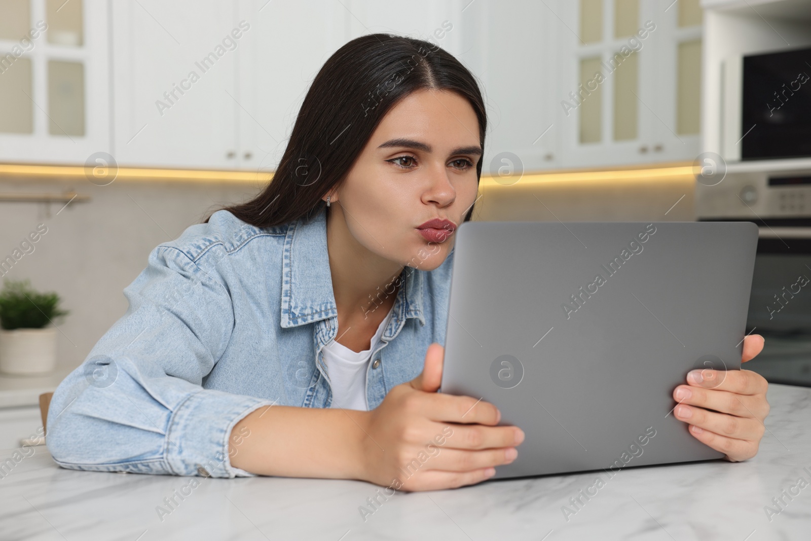 Photo of Young woman having video chat via laptop and sending air kiss at table in kitchen