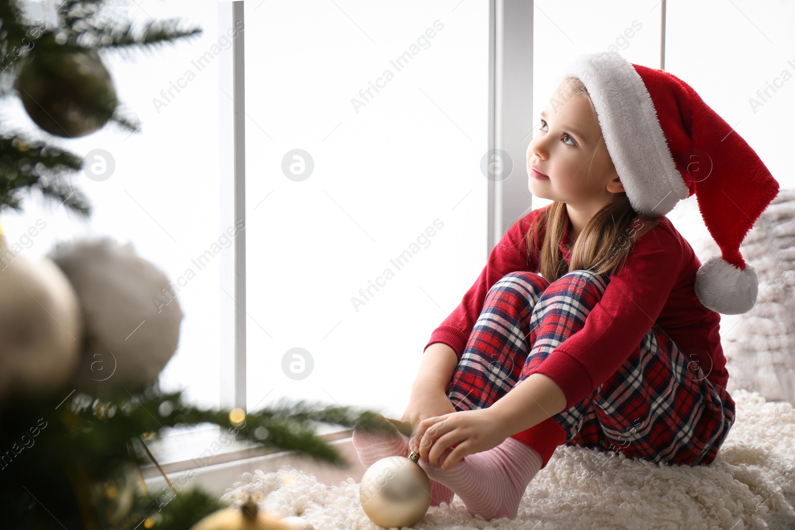 Photo of Cute little girl in Santa hat holding Christmas ball on window sill at home