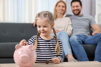 Family budget. Little girl putting coin into piggy bank while her parents watching indoors, selective focus