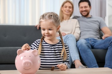 Photo of Family budget. Little girl putting coin into piggy bank while her parents watching indoors, selective focus