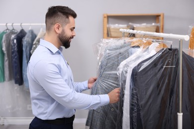 Photo of Dry-cleaning service. Happy worker choosing clothes from rack indoors