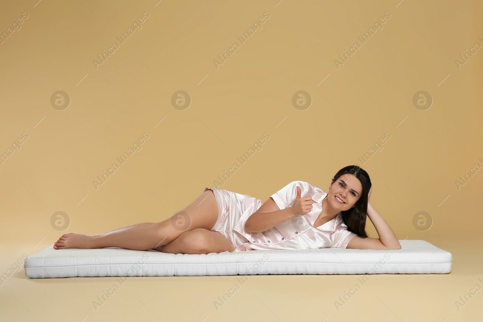 Photo of Young woman lying on soft mattress and showing thumbs up against beige background