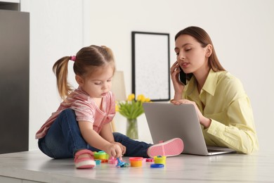 Photo of Woman working remotely at home. Busy mother watching over her daughter. Child sitting on desk