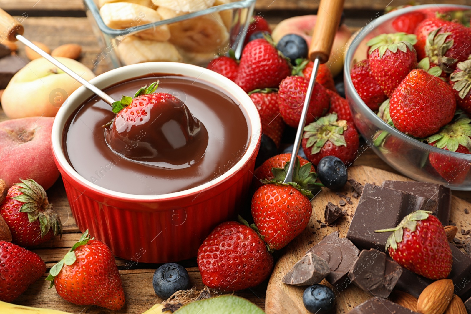 Photo of Fondue fork with strawberry in bowl of melted chocolate surrounded by other fruits on wooden table