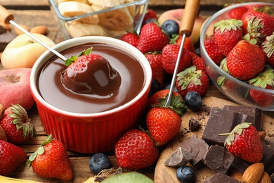 Photo of Fondue fork with strawberry in bowl of melted chocolate surrounded by other fruits on wooden table