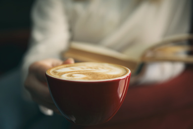 Woman with coffee reading book indoors, focus on cup