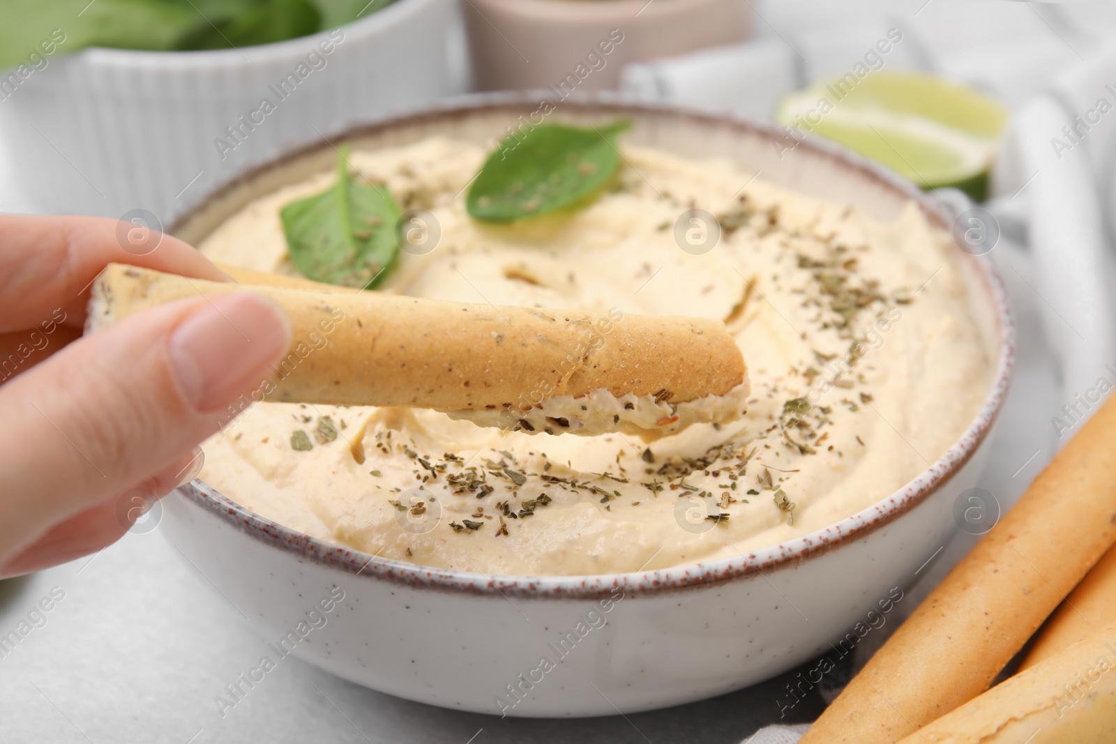 Photo of Woman dipping tasty grissini stick into hummus at light grey table, closeup