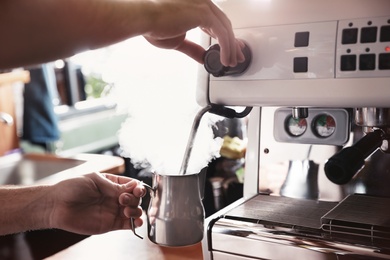 Photo of Barista steaming milk in metal jug with coffee machine wand at bar counter, closeup