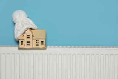 Photo of Modern radiator with knitted hat and wooden house near light blue wall indoors, space for text. Winter heating efficiency