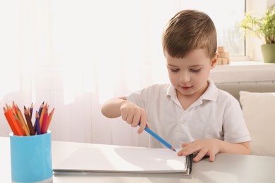 Photo of Cute little boy drawing with pencil at white table in room. Child`s art