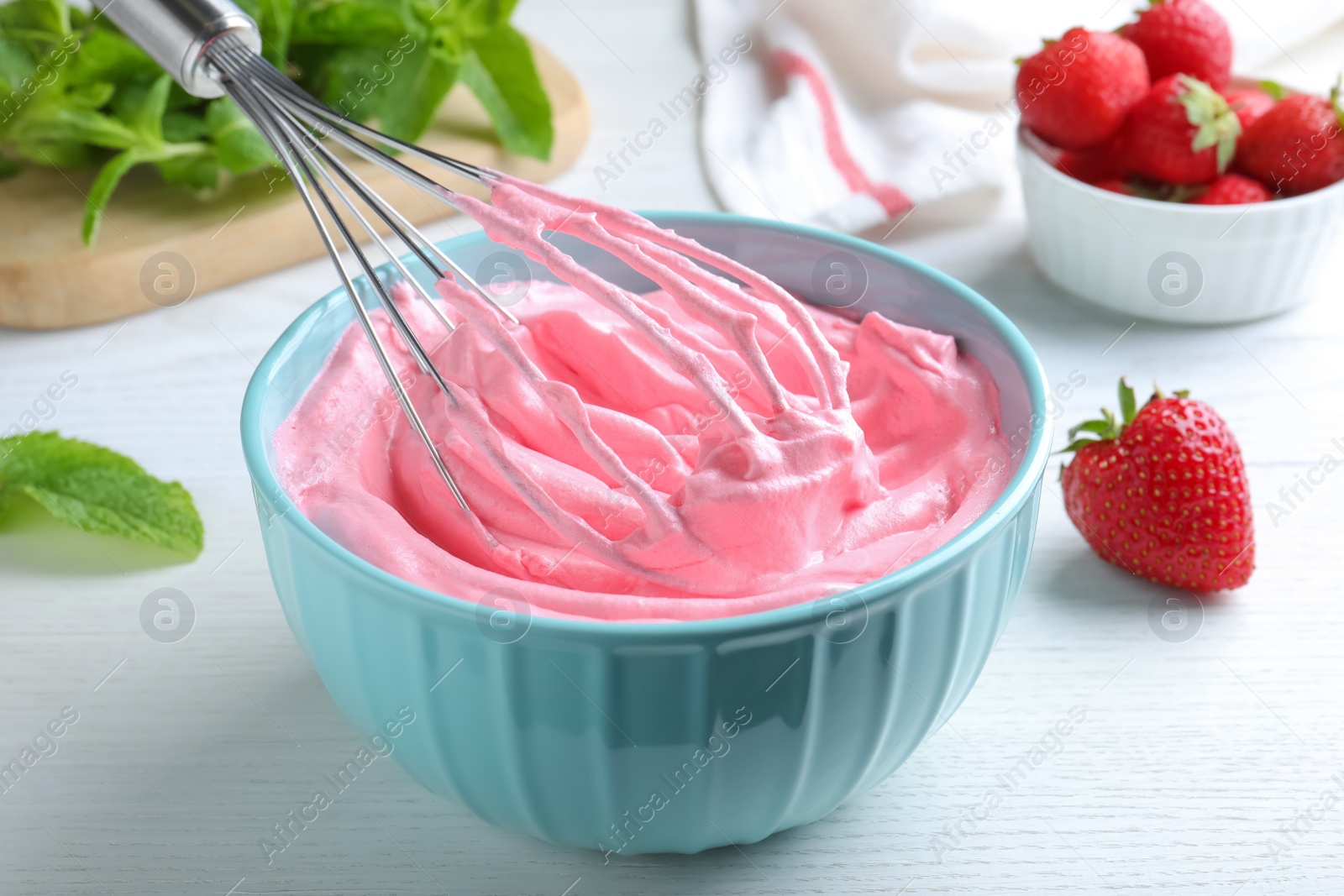 Photo of Whipping strawberry cream with balloon whisk on white wooden table, closeup