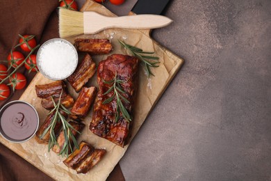 Photo of Tasty roasted pork ribs served with sauce, rosemary and tomatoes on brown table, top view. Space for text
