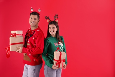 Beautiful happy couple in Christmas headbands and sweaters holding gifts on red background. Space for text