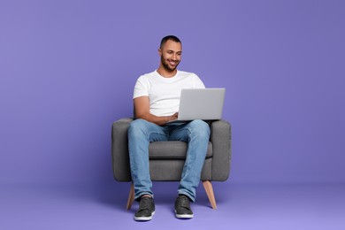 Photo of Smiling young man working with laptop in armchair on lilac background