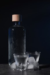 Photo of Bottle of vodka and shot glasses with ice on black table against dark background