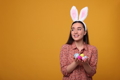 Photo of Happy woman in bunny ears headband holding painted Easter eggs on orange background. Space for text.