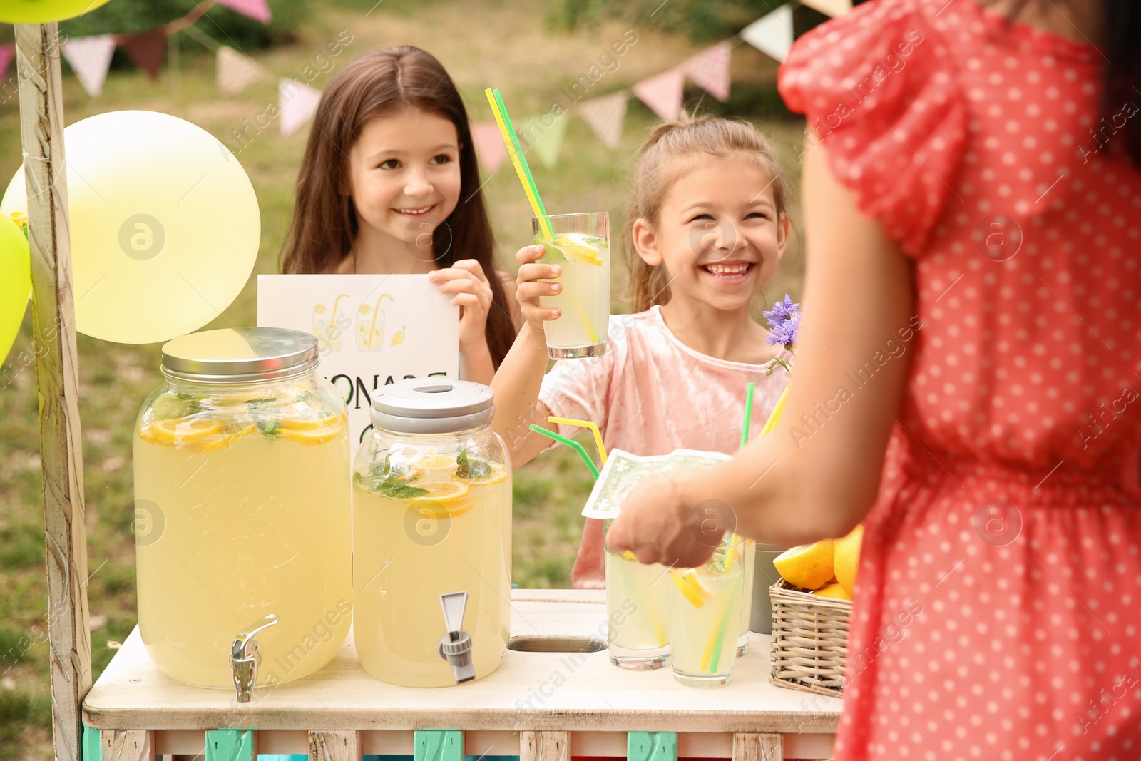 Photo of Little girls selling natural lemonade to woman at stand in park