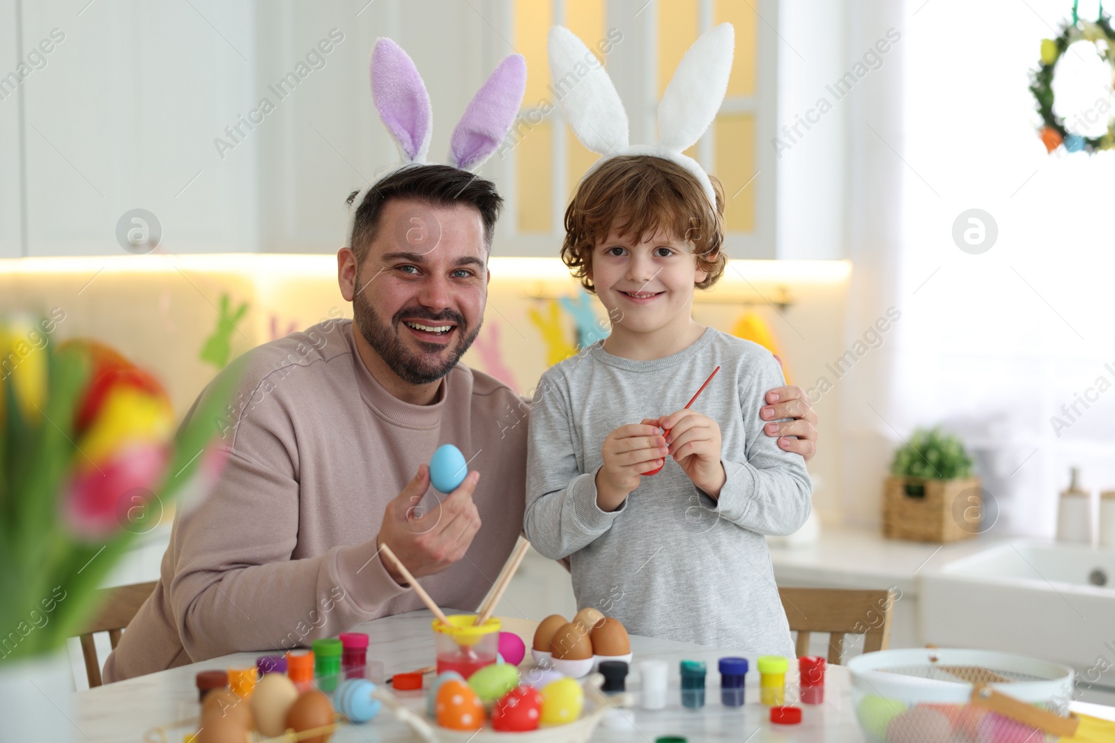 Photo of Easter celebration. Happy father with his little son painting eggs at white marble table in kitchen