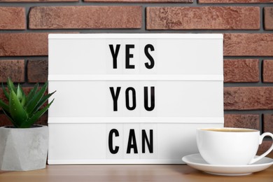 Photo of Lightbox with phrase Yes You Can, cup of coffee and potted houseplant on table against brick wall. Motivational quote