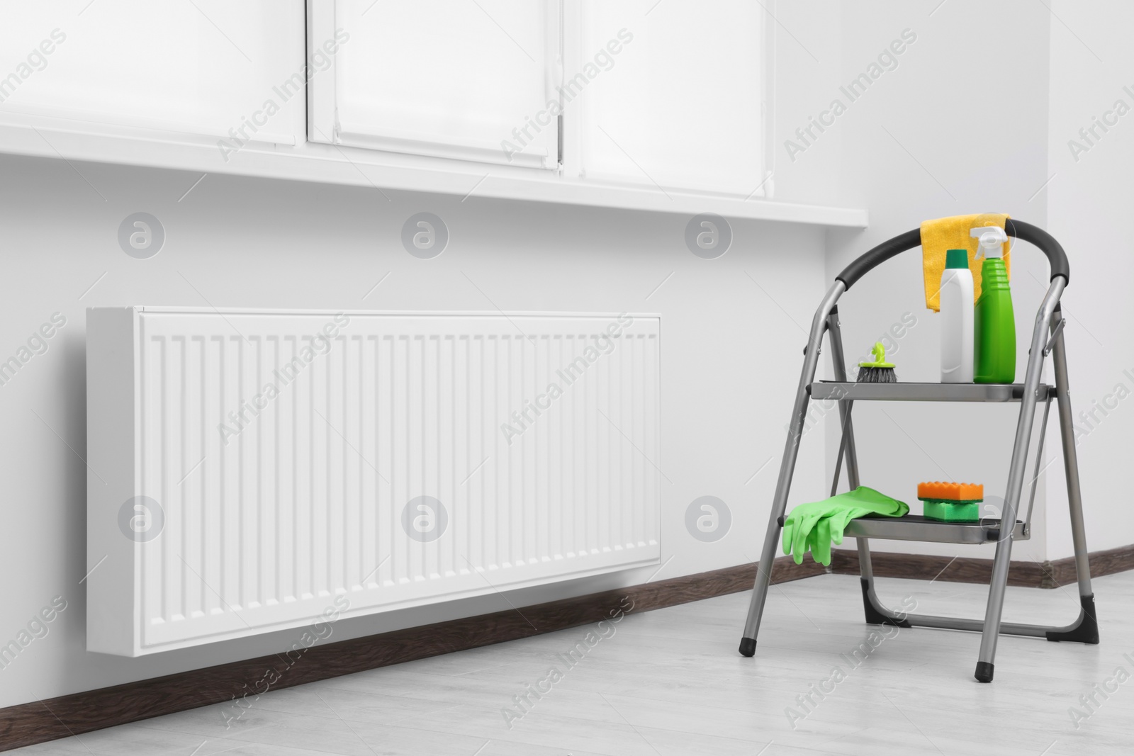 Photo of Ladder with cleaning supplies near modern radiator in room