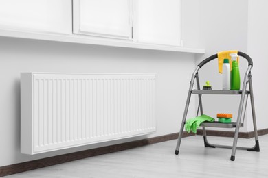 Photo of Ladder with cleaning supplies near modern radiator in room