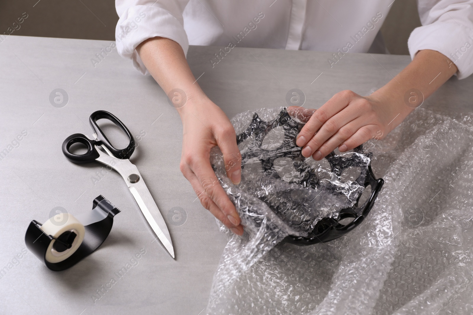 Photo of Woman covering ceramic bowl with bubble wrap at light grey table, closeup