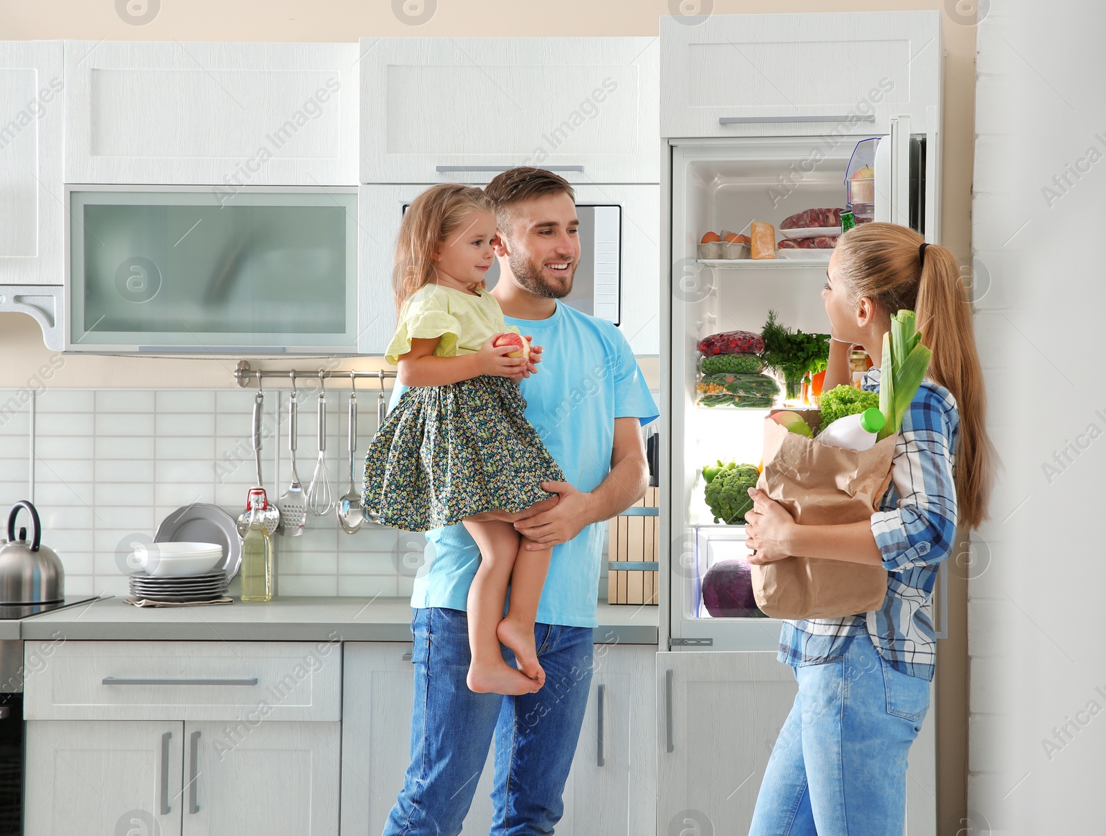 Photo of Happy family with paper bag full of products standing near refrigerator in kitchen