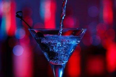 Pouring cocktail into martini glass with olives on blurred background, closeup