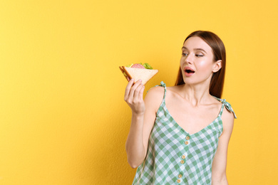 Young woman eating tasty sandwich on yellow background. Space for text