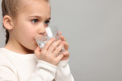 Cute little girl drinking fresh water from glass on light grey background, space for text