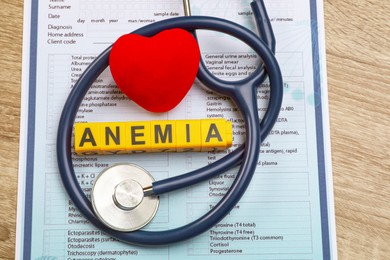 Photo of Word Anemia made with cubes, stethoscope, medical form and decorative heart on wooden table, above view