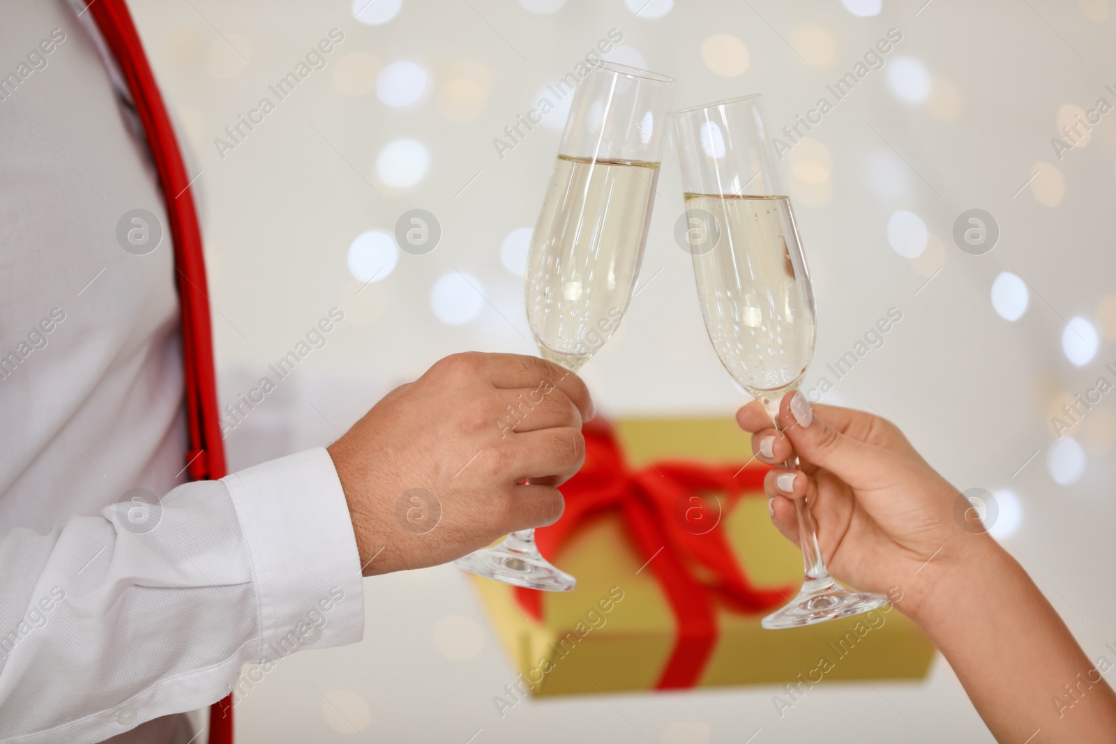 Photo of Young couple with glasses of champagne and gift box against blurred festive lights, closeup. Christmas celebration