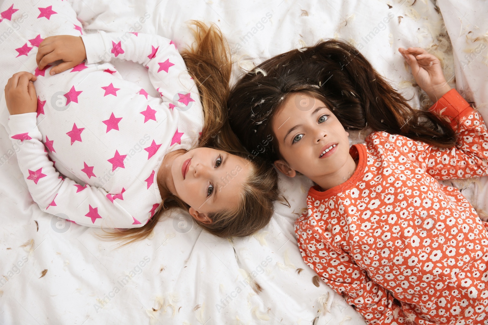 Photo of Cute little girls in pajamas lying among feathers on bed, top view. Happy childhood