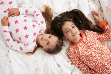 Cute little girls in pajamas lying among feathers on bed, top view. Happy childhood