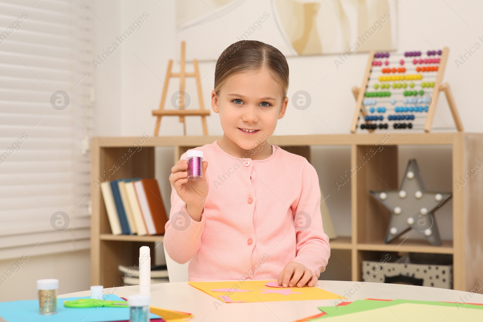 Photo of Cute little girl with container of colorful glitter at desk in room. Home workplace