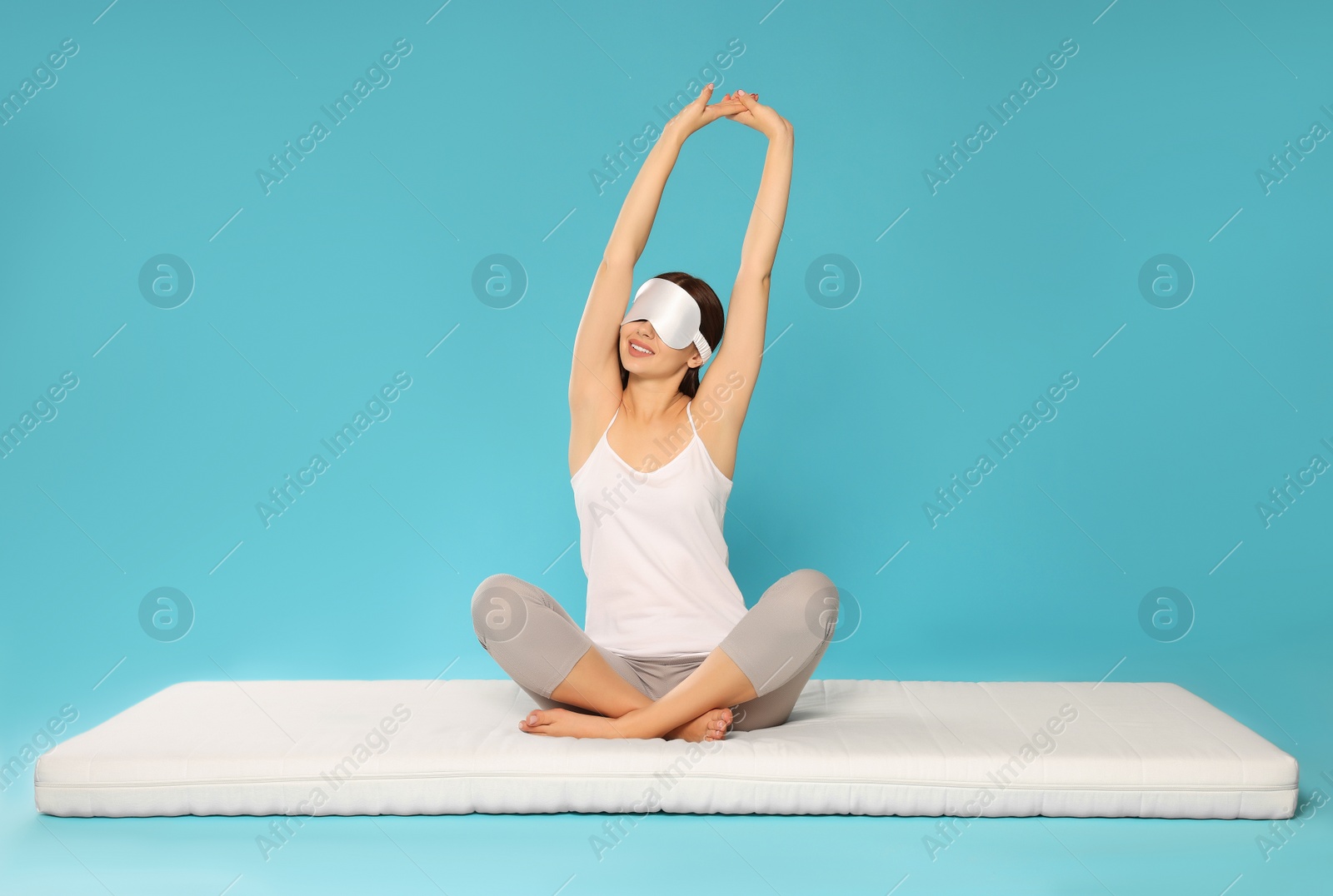 Photo of Woman in sleep mask stretching on soft mattress against light blue background