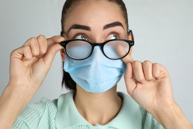 Photo of Woman wiping foggy glasses caused by wearing medical mask on light background, closeup